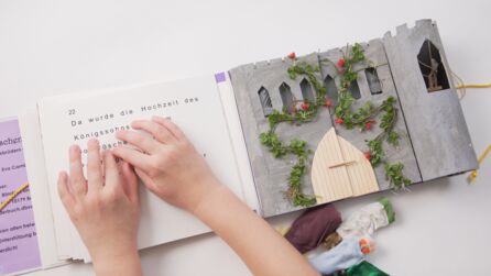 Kinderhände ertasten den Titel in Braille auf dem Tastbuch der Geschichte von Dornröschen. An der Seite des Buches ist ein ertastbarer Turm mit einer Öffnung angebracht. In der Öffnung ist eine kleine Figur mit einer Spindel.